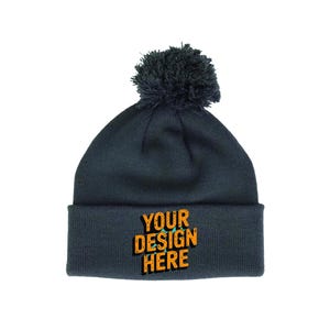 Könnte beinhalten: Dunkelgraue Strickmütze mit passendem Bommel oben. Der umgeschlagene Saum zeigt den Text "YOUR DESIGN HERE" in Orange und Gelb. Dieses Winteraccessoire ist stilvoll.