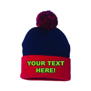 May include: A navy blue beanie hat with a red brim and a burgundy pom-pom. The red brim displays the text "YOUR TEXT HERE!" in green. This winter hat is a customizable accessory.