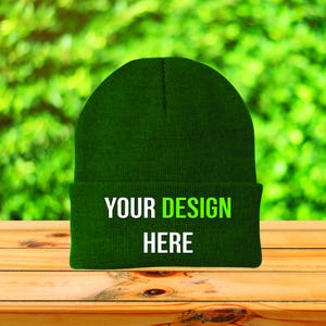May include: A dark green beanie hat with the text "YOUR DESIGN HERE" in white and green. The hat is displayed on a wooden surface with a blurred green background.
