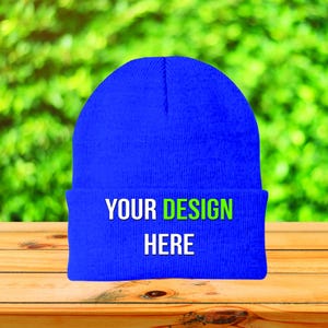 May include: A vibrant blue beanie hat with the text "YOUR DESIGN HERE" in white and green. The hat is made of a knitted material and is displayed on a wooden surface with a blurred green background.