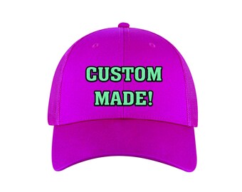 May include: A vibrant magenta baseball cap with a mesh back and a curved brim. The front of the cap features the words "CUSTOM MADE!" in green block letters with a black outline. The cap is designed for casual wear.