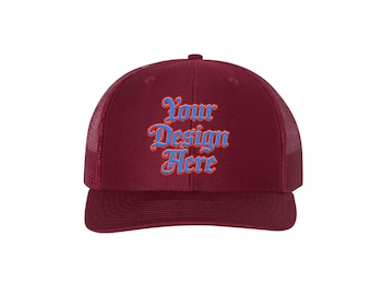 May include: Maroon trucker hat with a mesh back and a curved brim. The front of the hat features the text "Your Design Here" in a stylized font, with blue lettering outlined in orange.