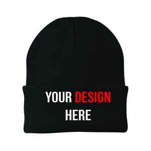 May include: A black knit beanie with a folded brim. The text "YOUR DESIGN HERE" is printed across the brim in white and red block letters. The beanie is a winter accessory.