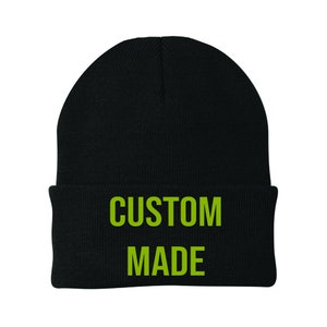 May include: A black beanie hat with the words "CUSTOM MADE" in bright green block letters. The hat is a classic knitted style, suitable for cooler temperatures.