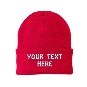 May include: A vibrant red beanie hat with a turned-up brim. The hat features white embroidered text that says "YOUR TEXT HERE". The hat is made of a knitted fabric and is set against a white backdrop.