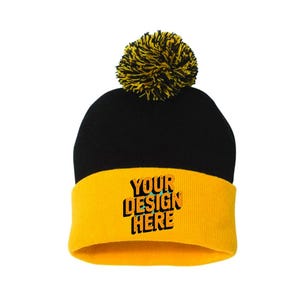 Puede incluir: Gorro de punto negro y amarillo con pompón. El gorro tiene un borde amarillo con el texto "YOUR DESIGN HERE" en una fuente multicolor. El pompón es amarillo y negro.