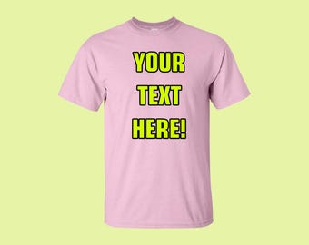 以下が含まれることがあります： 明るいピンク色のTシャツ。テキスト「YOUR TEXT HERE！」は、黒いアウトライン付きの明るい黄色の文字で表示されています。 Tシャツはクルーネックです。
