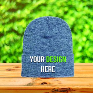 Peut inclure: Un bonnet bleu avec le texte "YOUR DESIGN HERE" en blanc et vert. Le chapeau est fait d'une matière douce et est présenté sur une surface en bois avec un arrière-plan vert flou.