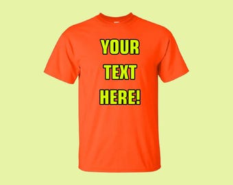以下が含まれることがあります： 鮮やかなオレンジ色のTシャツ。テキスト「YOUR TEXT HERE！」は、黒い縁取りのある太字の黄色で表示されています。Tシャツはクルーネックで、柔らかい素材で作られているようです。背景は明るい黄緑色です。
