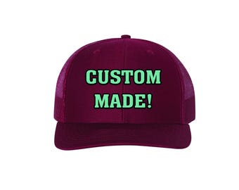 May include: A burgundy trucker cap with a mesh back and a curved peak. The front of the cap displays the words "CUSTOM MADE!" in a bold, mint green font. The cap is designed for casual wear.