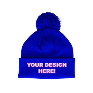 May include: A bright blue beanie hat with a fluffy pom-pom. The folded cuff features the text "YOUR DESIGN HERE!" in white with a pink outline. This winter hat is a customizable accessory.