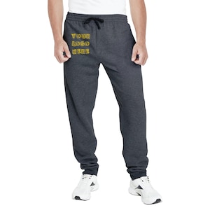 Puede incluir: Pantalones de chándal gris carbón con cintura con cordón y el texto "YOUR LOGO HERE" en amarillo. Los pantalones se combinan con zapatillas deportivas blancas. El estilo es informal y cómodo.