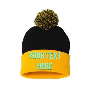 May include: A black beanie with a yellow brim and a yellow pom-pom. The brim features the text "YOUR TEXT HERE" in a turquoise color. The hat is set against a white background.