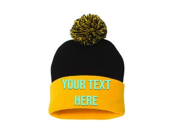 Può includere: Un berretto nero con bordo giallo e pompon giallo. Il bordo presenta il testo "YOUR TEXT HERE" in turchese. Il cappello è posto su uno sfondo bianco.