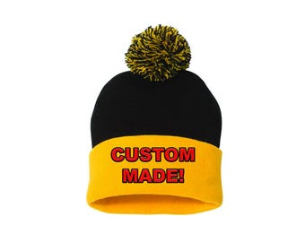 Può includere: Un berretto lavorato a maglia nero e giallo con un pompon giallo. Il berretto ha un bordo giallo con le parole "CUSTOM MADE!" in lettere rosse.