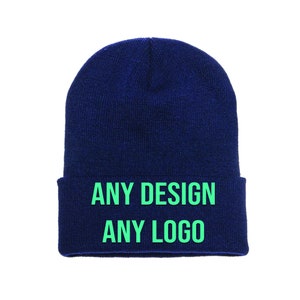May include: A navy blue beanie hat with a folded-up brim. The text "ANY DESIGN ANY LOGO" is printed in bright green across the folded brim. The hat is made of a soft, knitted material.