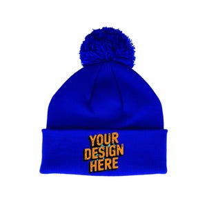 May include: A royal blue beanie hat with a matching pom-pom. The folded cuff displays the text "YOUR DESIGN HERE" in an orange, distressed font. This winter hat is a customisable accessory.