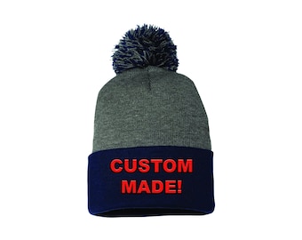 Può includere: Un berretto di maglia grigio con bordo blu navy. Il bordo presenta la scritta ricamata "CUSTOM MADE!" in rosso. Il cappello è sormontato da un pompon blu navy.