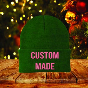 May include: A dark green beanie with the words "CUSTOM MADE" in pink block letters. The hat is made of a knitted material and has a folded brim. The background features a blurred Christmas tree with lights and ornaments.