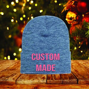 May include: A blue beanie with the words "CUSTOM MADE" in pink block letters. The hat is displayed on a wooden surface with a blurred background of Christmas lights and ornaments.