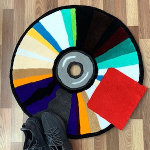 May include: A colorful circular rug with a red square in the lower right corner. The rug is designed to look like a vinyl record with a gray center and a black outer ring. Two black and gray sneakers are on the rug.