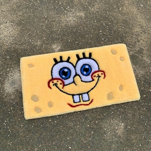 May include: A yellow bath mat with a cartoon face of Spongebob Squarepants. The face is embroidered with black eyelashes, blue eyes, a red smile, and white teeth.