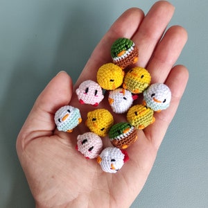 May include: A selection of small, crocheted chick ornaments in various colours, including yellow, white, pink, and green. Each chick has a detailed design with small beaks and eyes. The chicks are held in a hand against a teal background.
