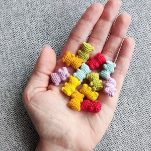Puede incluir: Una colección de pequeños osos de peluche de ganchillo en varios colores, incluyendo amarillo, rojo, verde y morado. Los osos se sostienen en una mano, mostrando sus detalles hechos a mano. El fondo es una superficie gris texturizada.