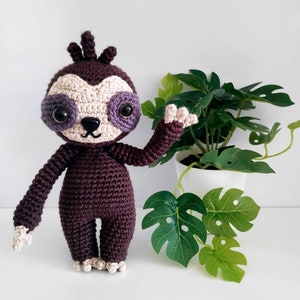 Puede incluir: Una muñeca de perezoso de ganchillo hecha a mano en tonos marrón, beige y morado. El perezoso tiene ojos grandes y expresivos y posa con un brazo levantado. Una maceta blanca con una planta de hojas verdes está en el fondo.