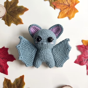 May include: A handmade crochet bat toy, featuring a grey body, large black eyes, and purple ear and wing accents. The bat is set against a white backdrop with autumnal leaves in red, orange, and yellow, and small wooden discs.