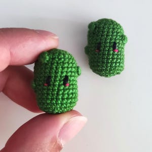 Puede incluir: Dos pequeñas criaturas verdes de ganchillo hechas a mano con ojos negros y bocas rosas. Una es sostenida por un dedo, la otra descansa sobre una superficie blanca. Las criaturas tienen pequeñas protuberancias en forma de oreja.