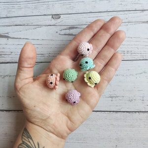 Puede incluir: Una colección de siete pulpos de ganchillo en miniatura en colores pastel: rosa, azul claro, verde, amarillo y morado. Cada pulpo tiene pequeños ojos negros. Los pulpos se muestran sobre una superficie de madera clara.