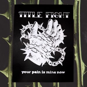 以下が含まれることがあります： 上部に「TITLE FIGHT」の文字が入った白黒のグラフィックプリント。 棘のある鎖に囲まれた手が描かれています。 下部には「your pain is mine now」の文字が入っています。