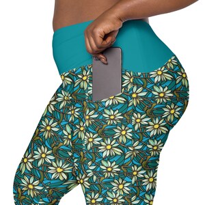May include: Teal leggings with a floral pattern of white daisies on a blue background. The leggings have a large pocket on the right side, with a black smartphone inside.