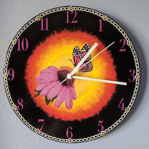 May include: A black clock with a painted design of a butterfly and flower. The clock face has white hands and pink numbers. The background is a gradient of orange, yellow, and red.
