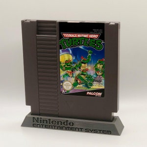 May include: A grey Nintendo Entertainment System game cartridge for Teenage Mutant Hero Turtles. The cartridge is on a grey stand with the words "Nintendo Entertainment System" printed on it. The game cartridge has a black label with the title "Teenage Mutant Hero Turtles" in white text. The label also features a colourful image of four Teenage Mutant Ninja Turtles.