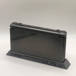 May include: A black Nintendo 3DS XL console on a black stand. The console has the text "new Nintendo 3DS XL" printed on the front.