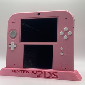 May include: A pink Nintendo 2DS handheld gaming console with white buttons and a stand. The console has two screens, one on top and one on the bottom.
