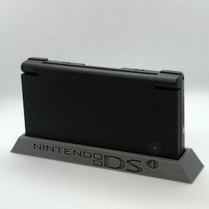 May include: A black Nintendo DS handheld gaming console in a grey 3D printed stand with the words "Nintendo DS" printed on the front.