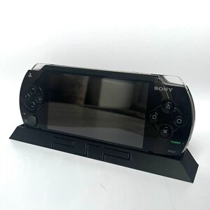 May include: Black Sony PSP handheld games console with a clear protective cover, on a black charging stand. The console has a screen, directional pad, buttons, and the Sony logo. Power and hold buttons are visible.