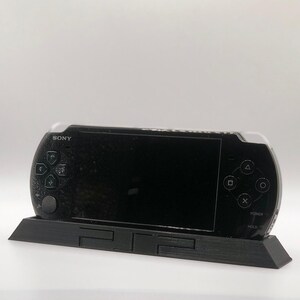 May include: A black Sony PlayStation Portable (PSP) handheld gaming console is sitting on a black stand with the PlayStation logo. The PSP has a black screen and buttons.