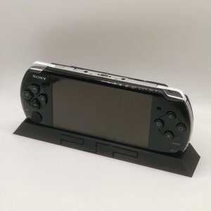 May include: A black Sony PlayStation Portable (PSP) handheld gaming console is sitting on a black stand. The PSP has a black and white design and features a screen, buttons, and a directional pad. The stand has a PlayStation logo on it.