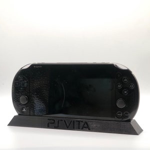 May include: A black PlayStation Vita handheld gaming console on a black stand with the words "PS VITA" printed on the front.