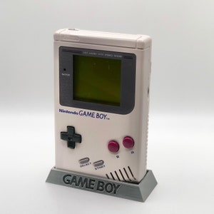 May include: A white Nintendo Game Boy handheld gaming console with a grey stand. The console has a black and grey control pad, two pink buttons, and a black and grey screen with the text "DOT MATRIX WITH STEREO SOUND". The stand has the text "GAME BOY" printed on the front.