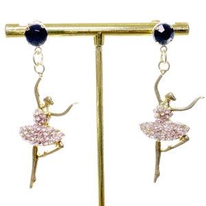 May include: A pair of gold-tone earrings featuring a ballerina silhouette adorned with pink rhinestones. Each earring has a black crystal at the top.