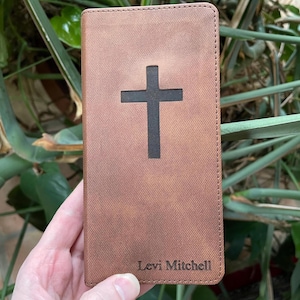 May include: A brown leather Bible cover with a black cross cutout. The cover has a textured appearance and the name "Levi Mitchell" is printed at the bottom. The cover is rectangular and appears to be new.