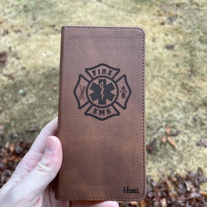May include: Brown leather phone case with a debossed Fire EMS emblem and the name "Hunt" at the bottom. The case has a textured surface and a stitched border. The emblem includes a Maltese cross, a star of life, and a fire hydrant.