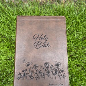 May include: Brown leather-bound Holy Bible with floral and butterfly design. The cover features the words "Holy Bible" in elegant script, along with the name "Brenda Ann" at the bottom. The Bible is set against a backdrop of green grass.