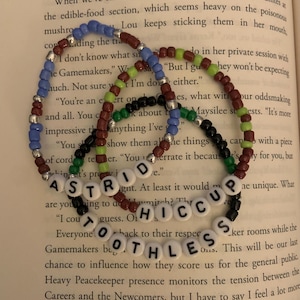 May include: Four beaded bracelets with letter beads spelling out the names "Astrid", "Hiccup", "Toothless" and "Otherless". The bracelets are made with colorful beads in shades of blue, green, red, and black.