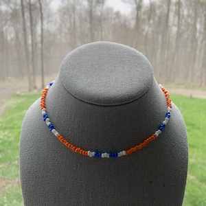 May include: A beaded necklace with orange, white, and blue beads. The necklace is on a gray mannequin head.